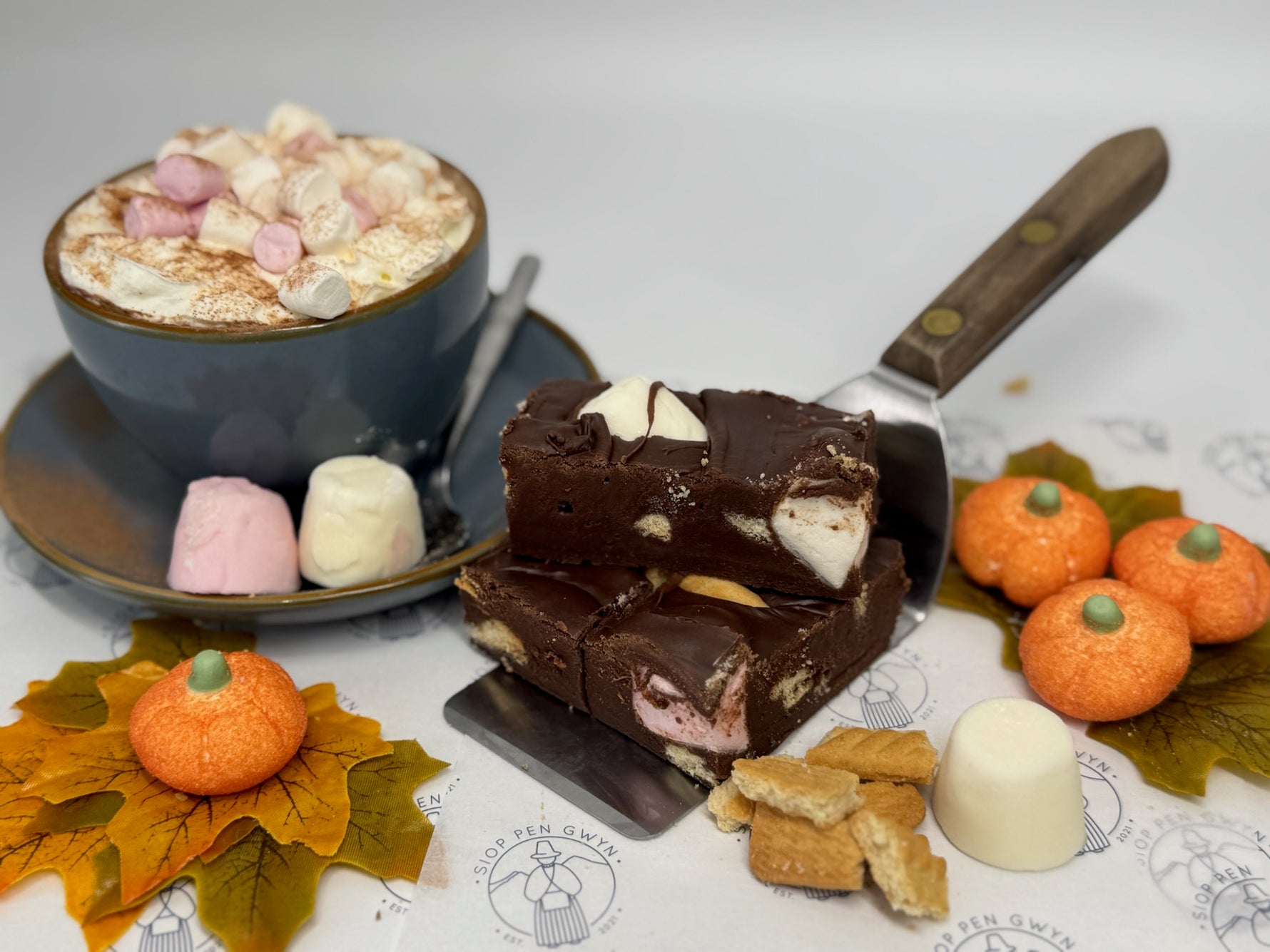 Chocolate dessert with marshmallows on a spatula, hot chocolate with marshmallows, and decorative pumpkins on a table.