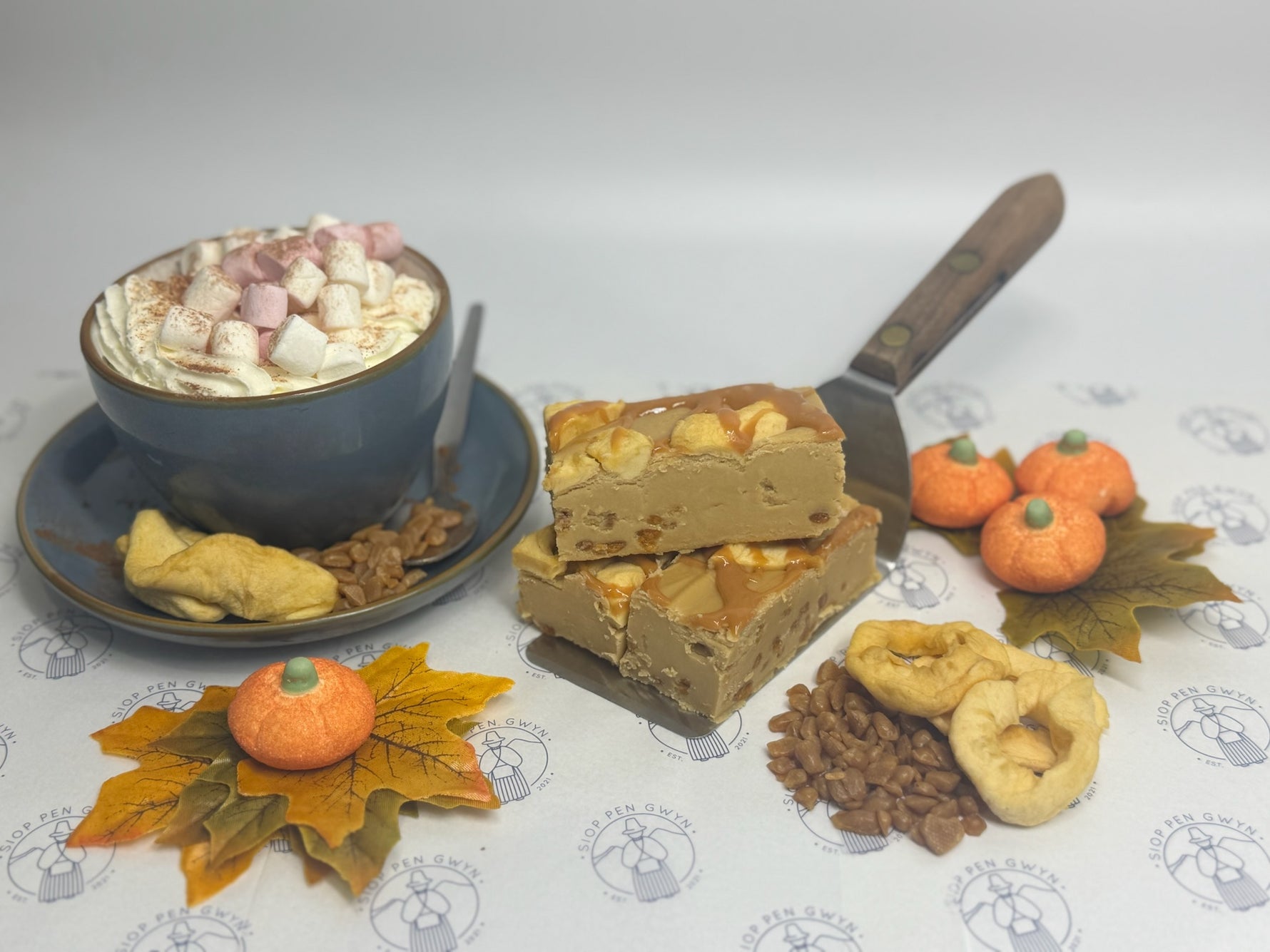 Dessert platter with a bowl of hot chocolate, slices of cake, and decorative pumpkins on a patterned tablecloth.