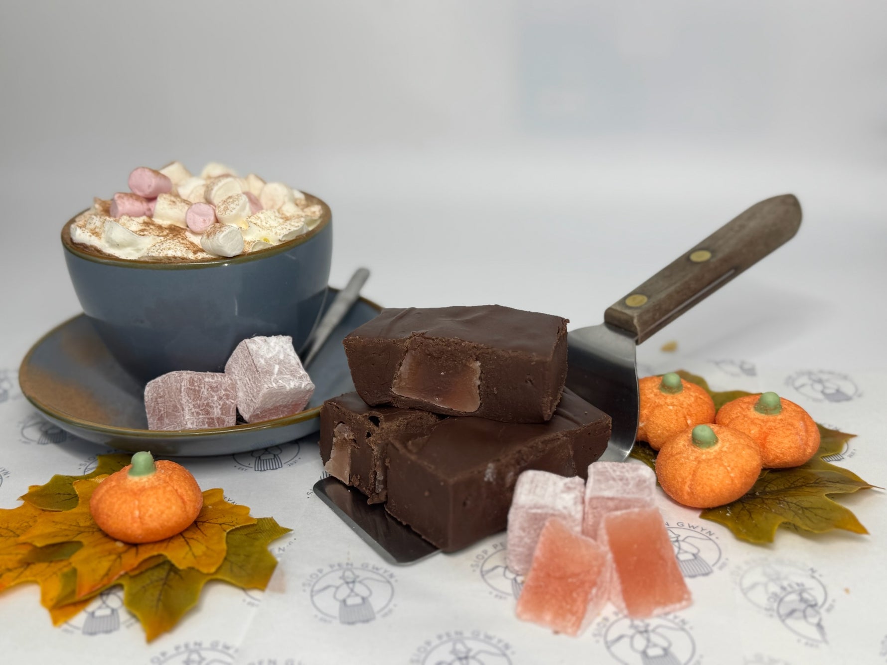 Chocolate fudge, marshmallows, and small pumpkins on a decorative tablecloth.