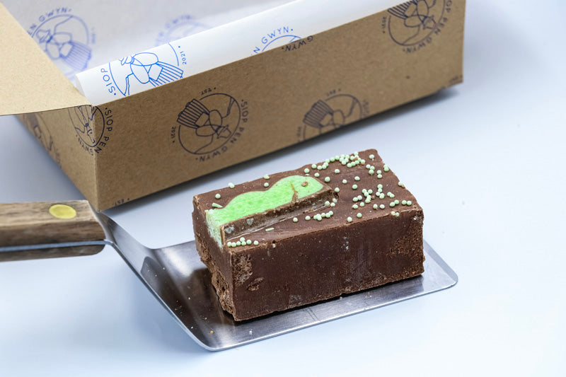 A piece of mint chocolate fudge with a swirl of green mint icing on top, placed on a silver platter beside a wooden spatula.